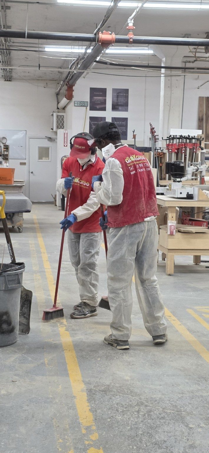 Deux préposés DIKAROO en tenue de protection balaient le plancher de béton d'un atelier.